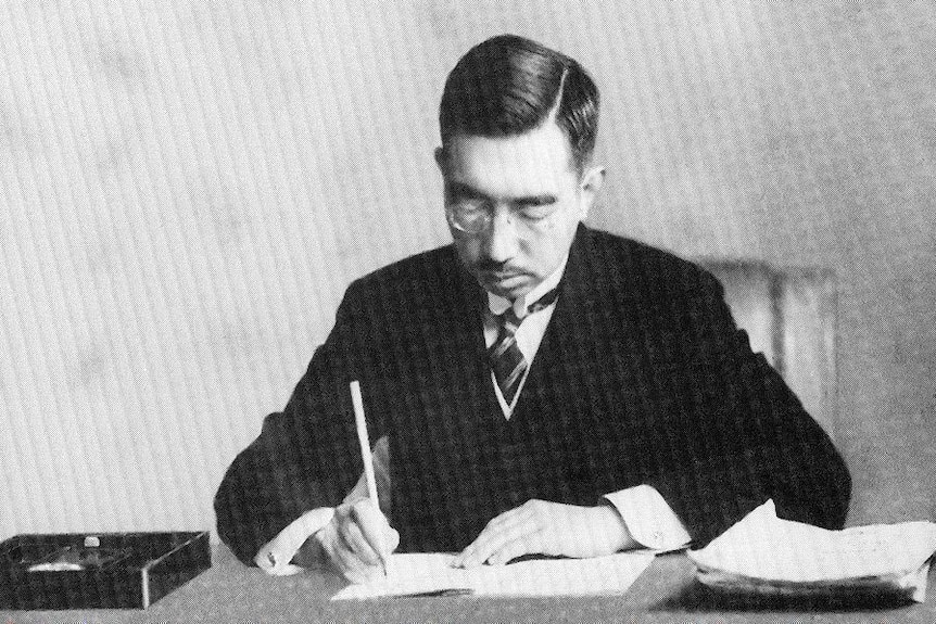 A black and white photo shows Emperor Hirohito sitting at a desk signing a new constitution into law.