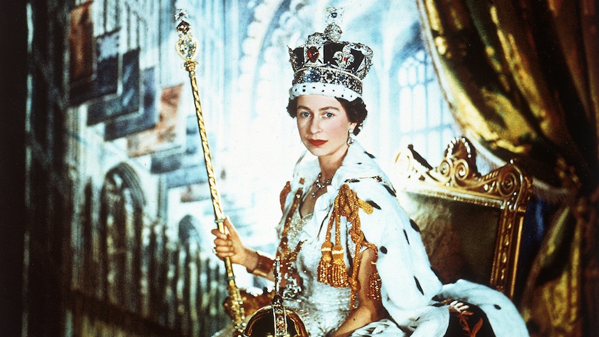 Queen Elizabeth II sitting in an ornate chair, wearing a heavy crown and carrying orb and sceptre.