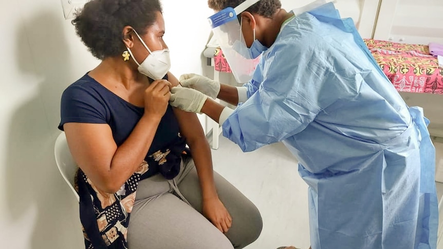 A woman sits in a chair wearing a mask and holding her arm as someone in scrubs and wearing a mask bends over her arm.