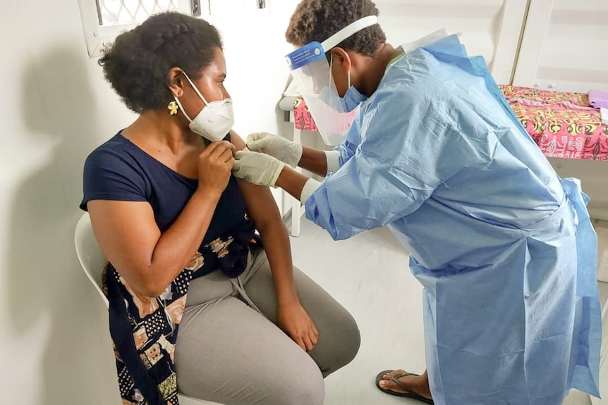 A woman sits in a chair wearing a mask and holding her arm as someone in scrubs and wearing a mask bends over her arm.