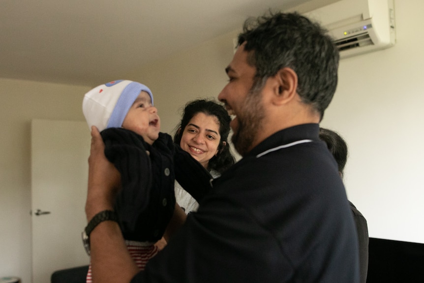 An Indian man holds a smiling baby while a woman looks on.