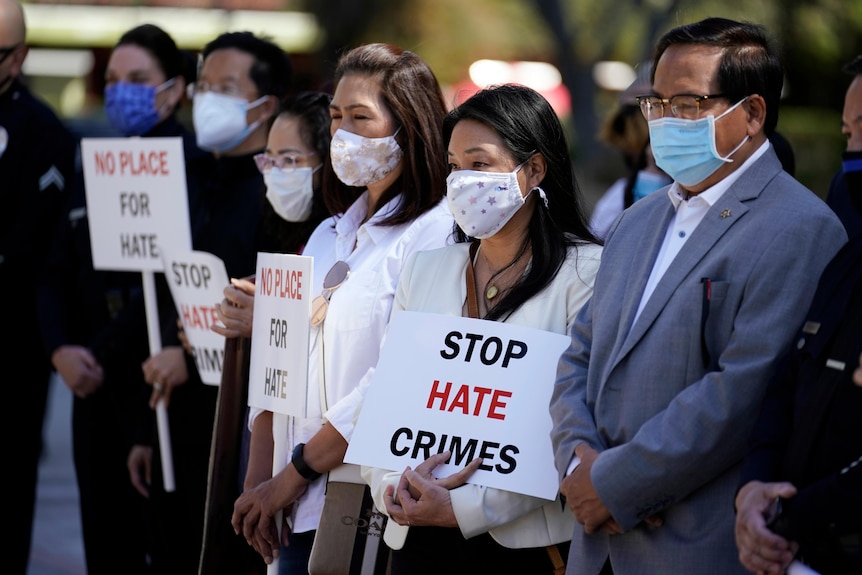 A group of face-masked Asians hold Stop Hate Crimes signs in a line on a sunny day.