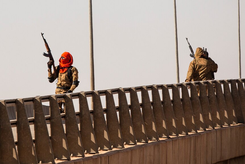 Two men with long arms in military uniforms and with faces covered walk across a bridge.