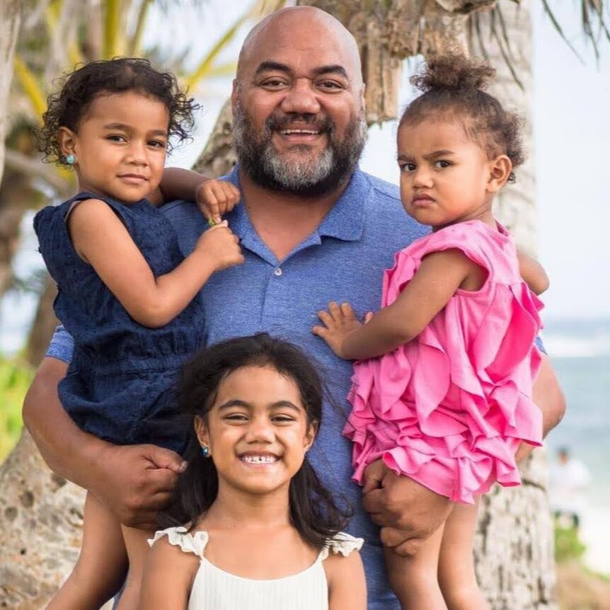 A tongan man standing on the beach holding two children besides another two children