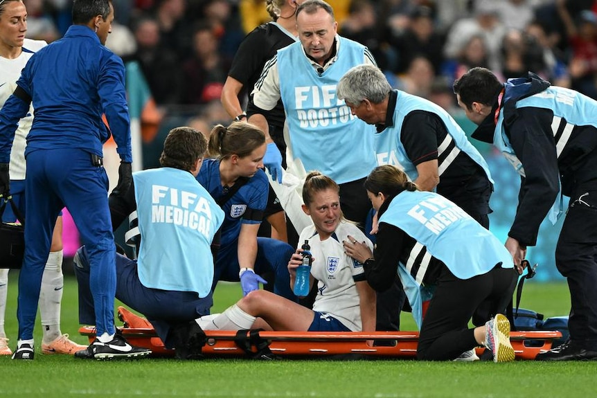England female football player is surrounded by medics, while sitting on a stretcher