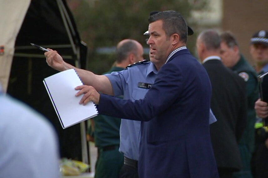 A man in a dark blue suit stands holding a notepad and talking to a WA police officer near other police and emergency services.