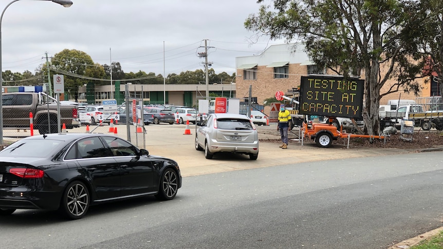 A sign reading 'testing site at capacity' with two cars in the driveway 