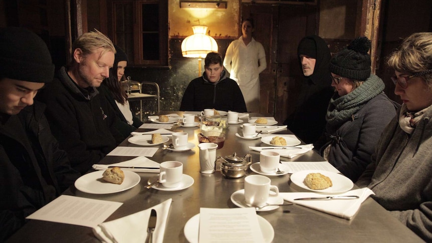 7 people sit at a table, each has scone on plate in front of them. Table set with tea. Performer in white uniform in background.