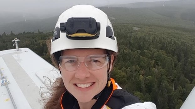 A woman in a white hardhat, clear goggles and a white work suit stands on a wind turbine high about a forest