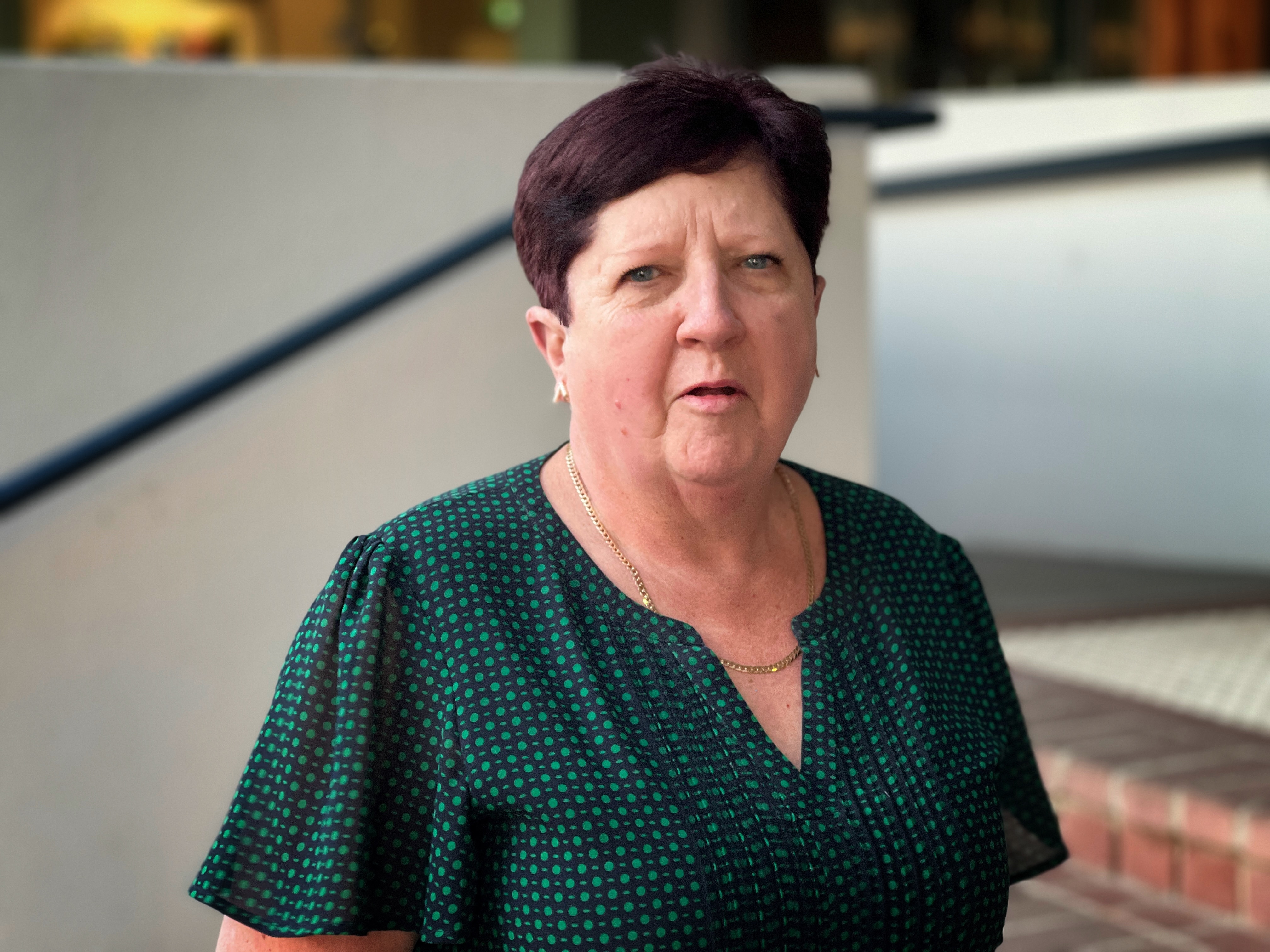 Carol Sandland in a green shirt, looking straight at the camera out the front of a building. 