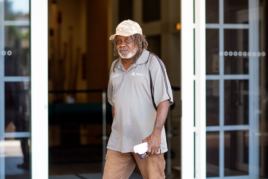 A man with dark skin and a grey beard wearing a cap leaving court