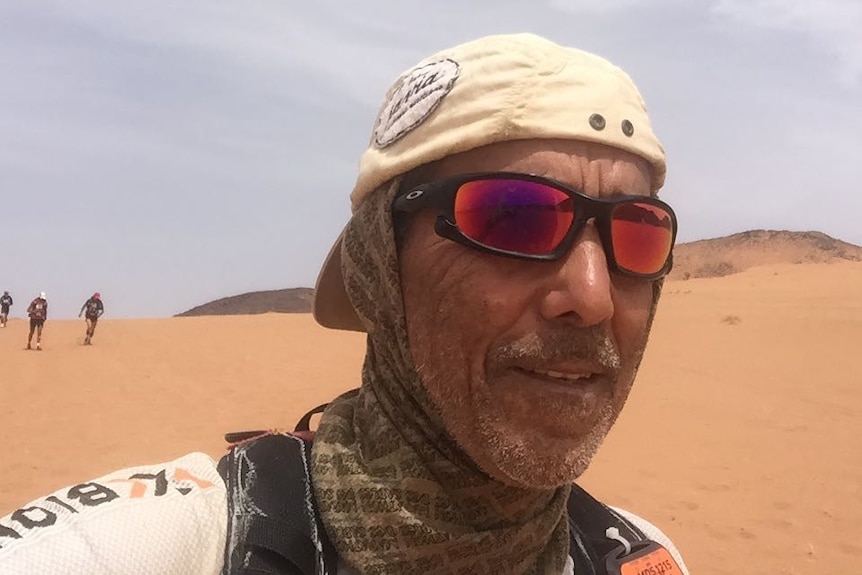 Un homme portant des lunettes de soleil, un chapeau et une écharpe enroulée autour du cou se tient sur une dune de sable