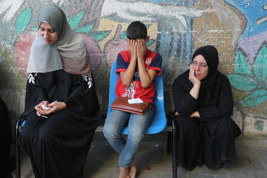Three people sitting by a wall, crying