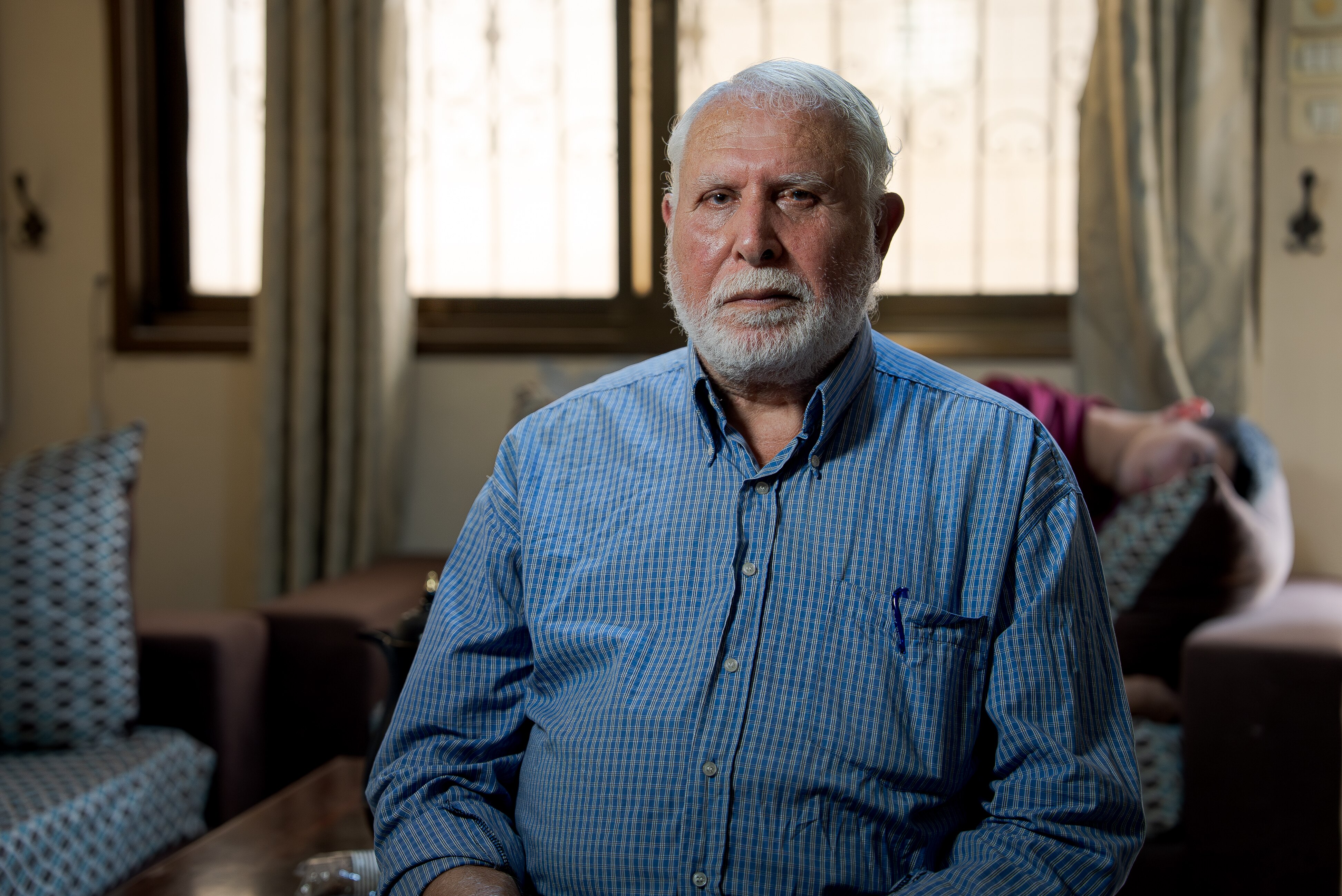 A man with white hair and a white beard sits in a chair with a blue shirt on.