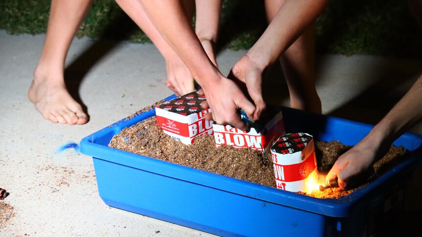 Two people leaning down and lighting several small fireworks arranged in a box filled with sand, on the ground.