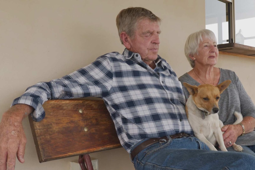 Ron and Ellen Stewart sitting next to each other on a bench seat on a verandah with a small dog sitting across their laps