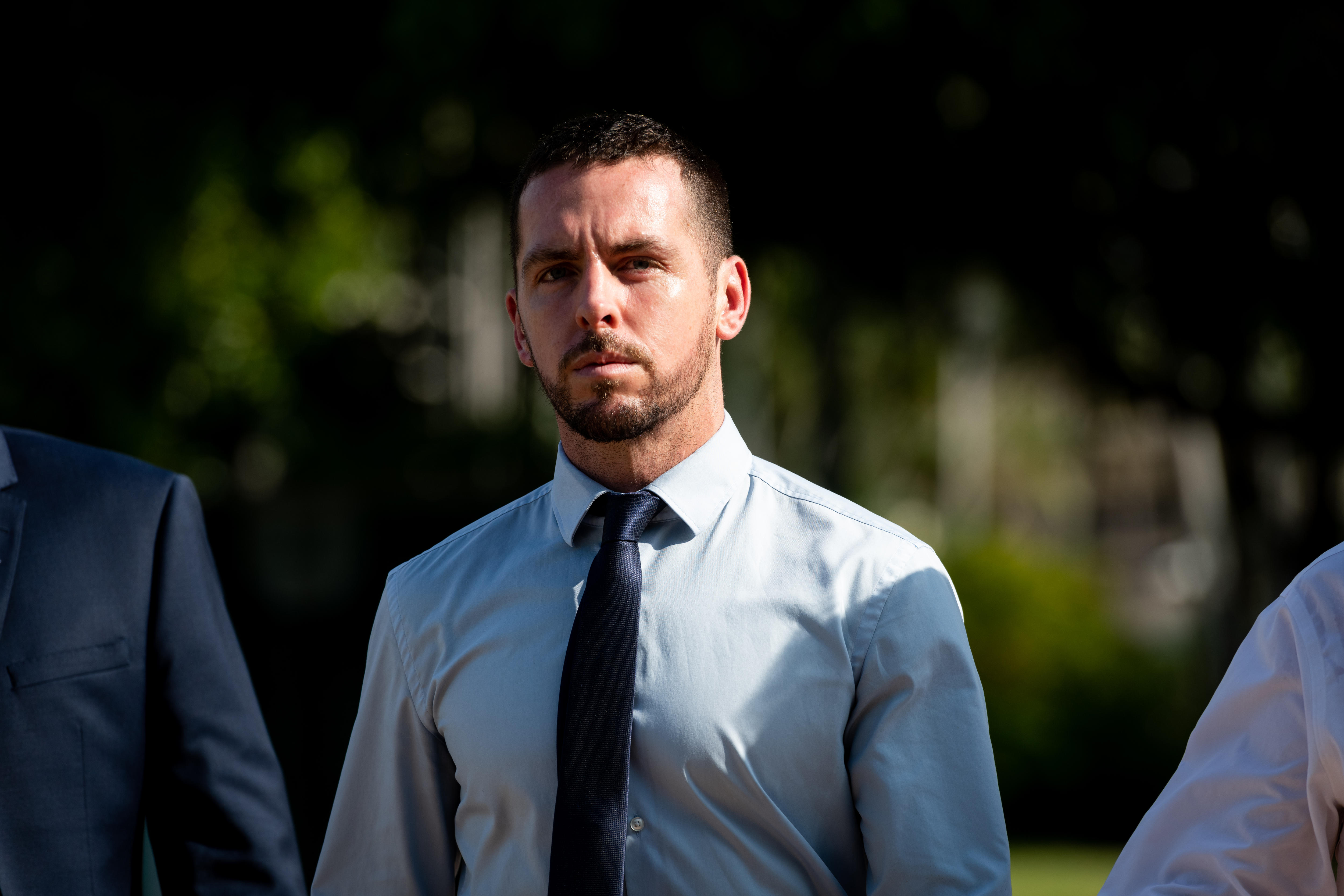 A man wearing a pale blue business shirt and navy tie walks with a stern expression.