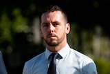 A man wearing a pale blue business shirt and navy tie walks with a stern expression.