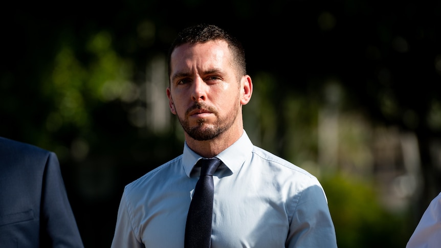 A man wearing a pale blue business shirt and navy tie walks with a stern expression.