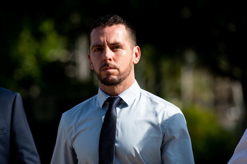 A man wearing a pale blue business shirt and navy tie walks with a stern expression.