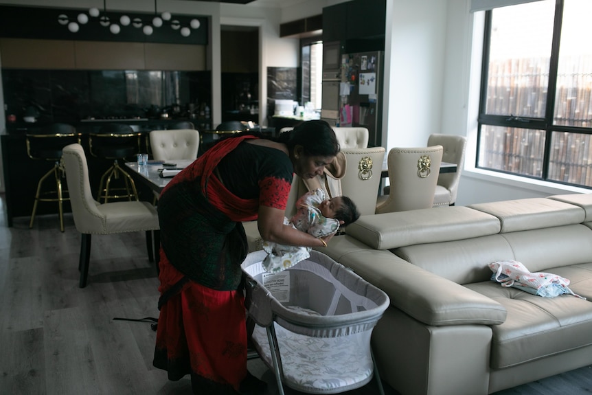 A woman in a black and red saree picks baby up from crib in a room with cream leather couches and leather chairs.