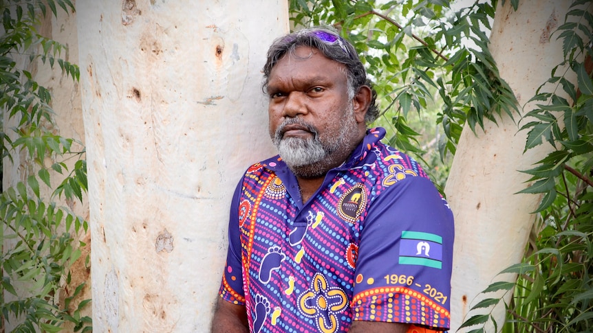 Middle aged Indigenous man with colourful shirt, sunglasses on top of head and beard, stands in front of gum tree