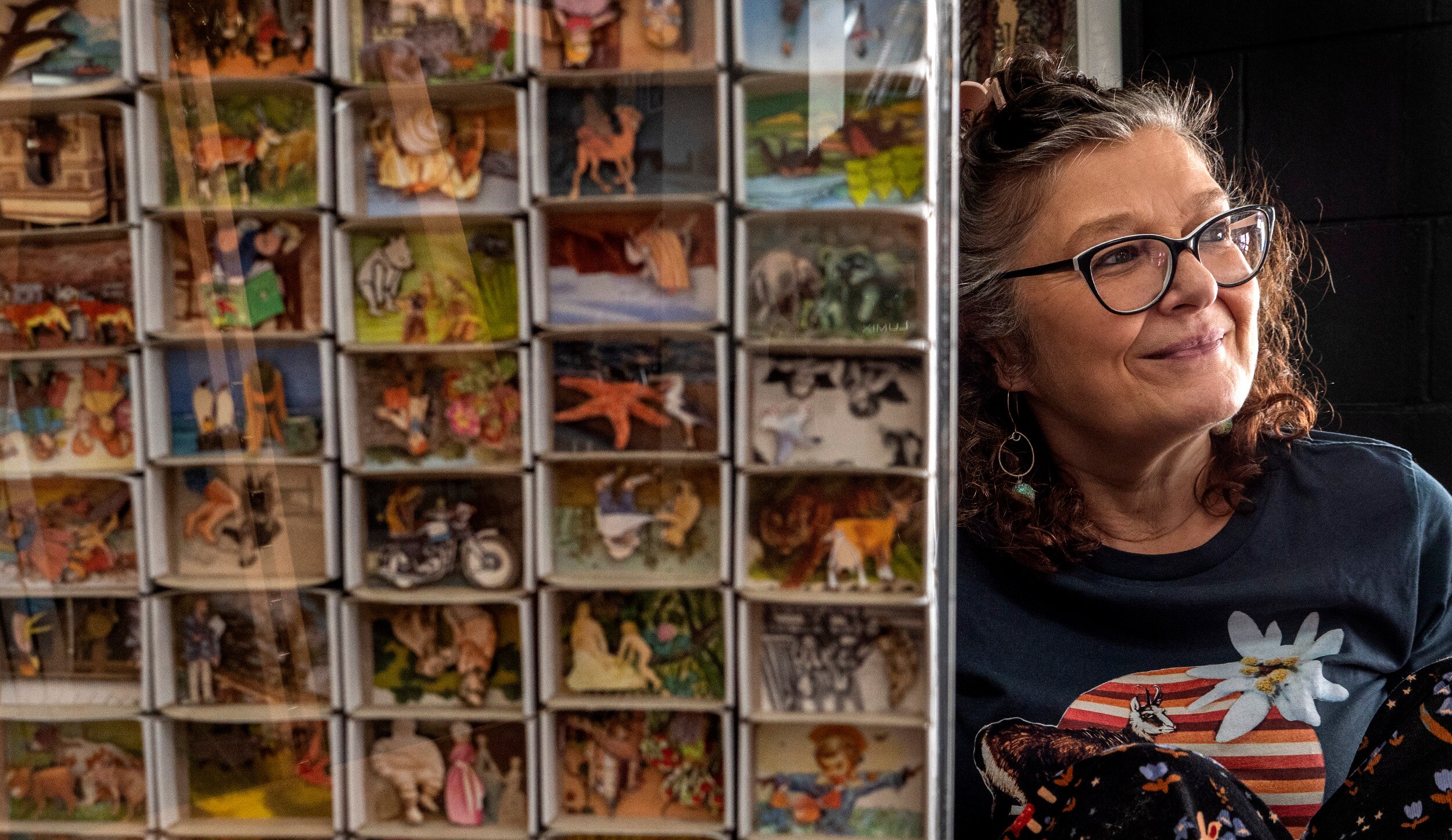 A woman leans against an artwork of empty matchboxes filled with collage stories.
