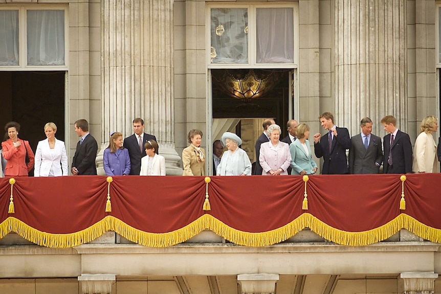 A group of royals dressed in formal attire stand on a blacony decorated in red curtain with a yellow trim.
