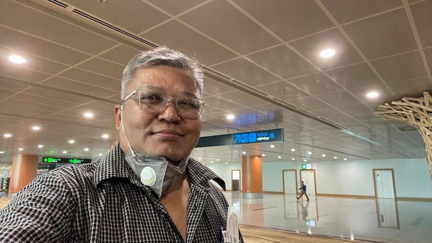 A selfie of a man with a mask pulled down and airport signs in the background.