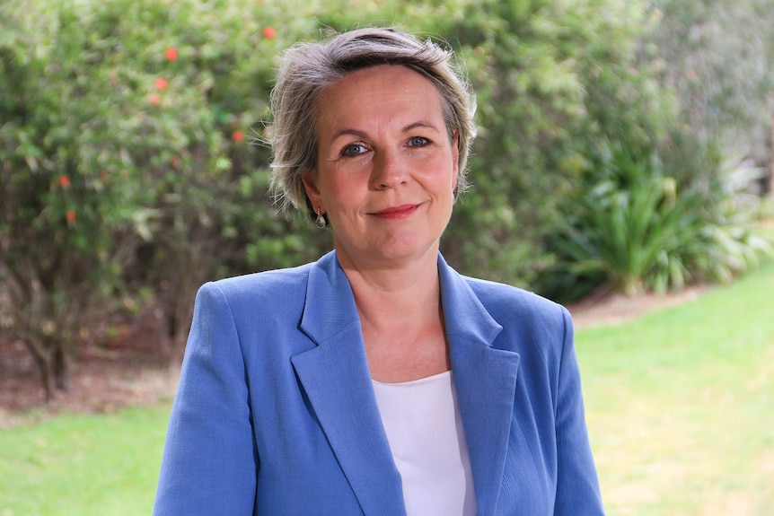 A fair-haired woman wearing a white top and pale blue blazer looks directly into the camera as she stands near a garden