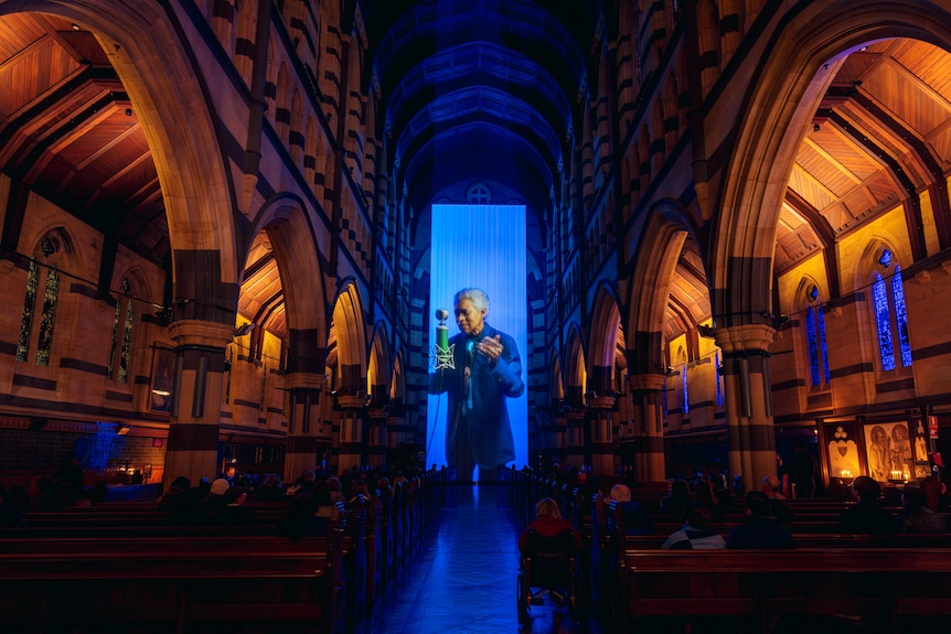 A view down the aisle of a cathedral, awash in golden light. At the end of the aisle, a singer is projected onto a blue screen.