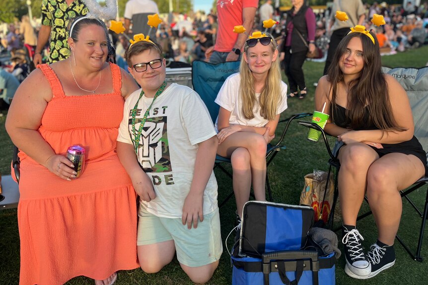 A medium shot of a woman and three young people sitting among a crowd outdoors with drinks and food, smiling.