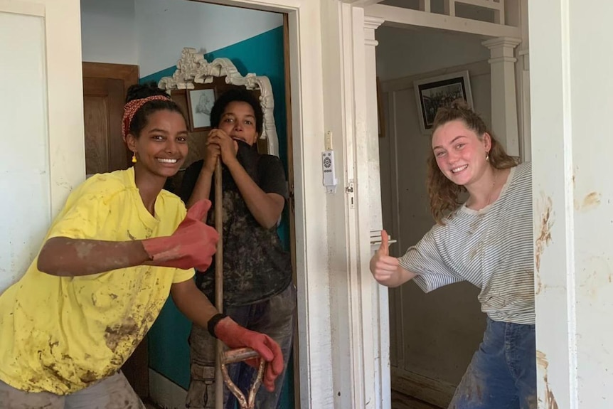 Three girls hold their thumbs up at the camera as they stand with cleaning supplies in a flood-ravaged home.