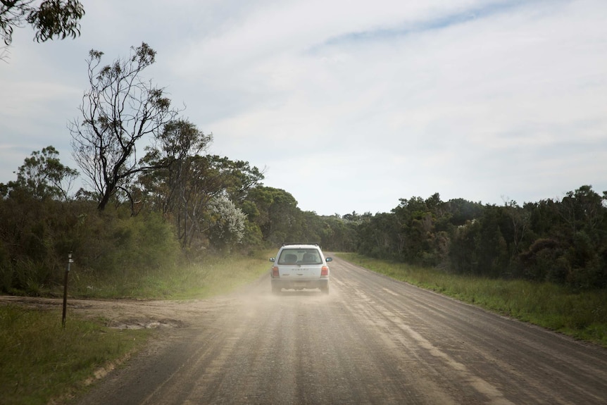 A car throws up dust as it drives along a dirt road, framed by scrub.