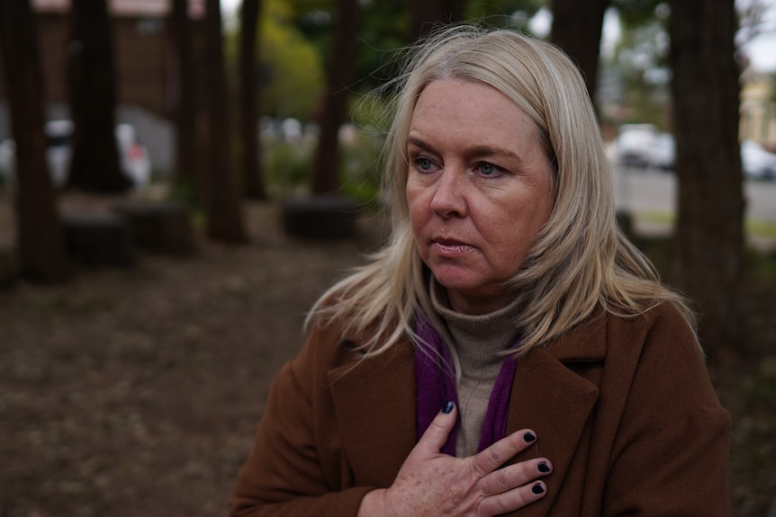 A woman stands outside with her hand on her chest.