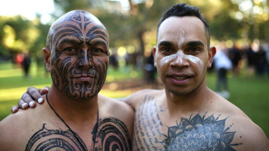 A young Indigenous man in white face paint embraces a man with Maori head tattoos