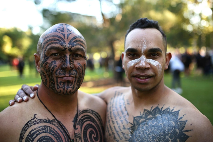 A young Indigenous man in white face paint embraces a man with Maori head tattoos