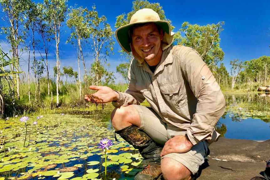 A man squats in front of a pond holding an unknown water species