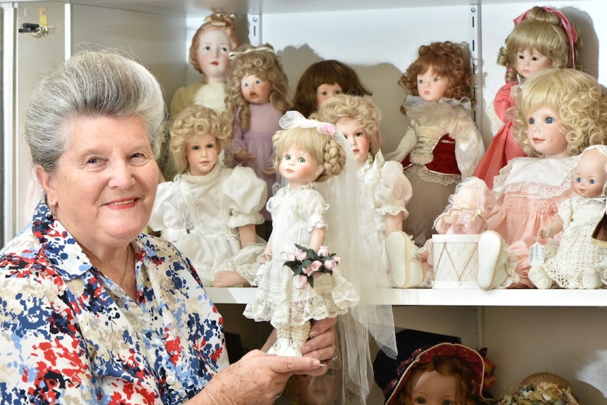 Woman with grey hair standing in front of a cabinet of porcelain dolls