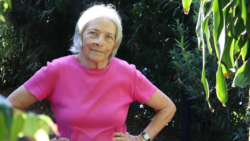 An elderly woman stands in a garden surrounded by trees with her hands on her hips and looking at the camera.