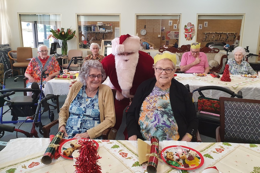 A man dressed as Santa stands behind two seated elderly women with other elderly men and women in the background.