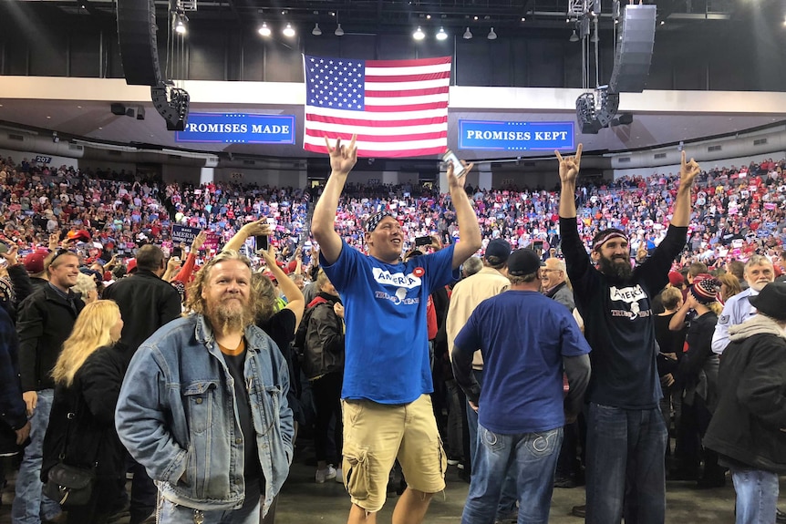 Three men pose in front of a large crowd inside an arena decorated with a large American flag and campaign banners