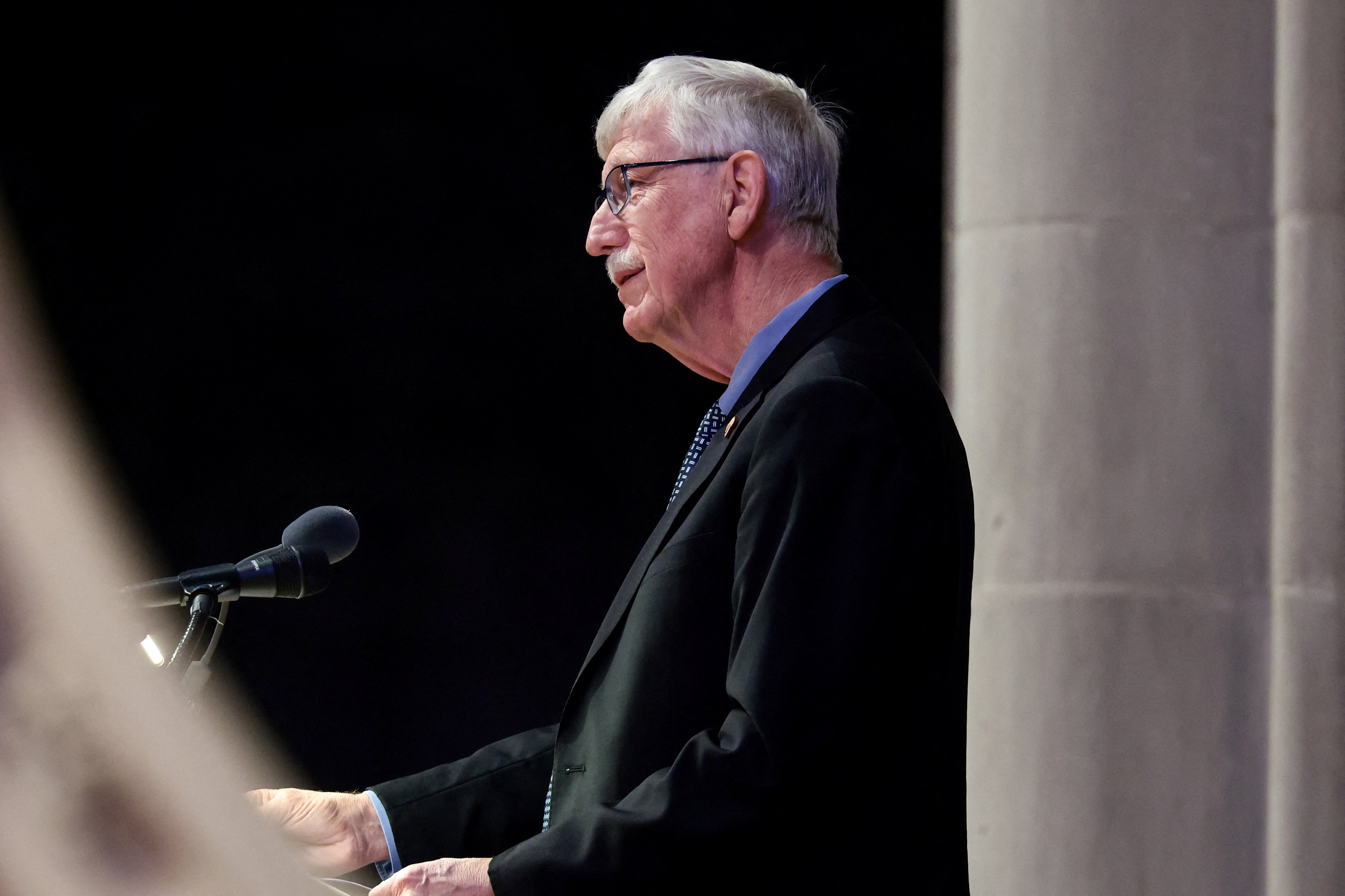 A grey-haired man in a black suit speaks into a microphone at a lectern in a cathedral.