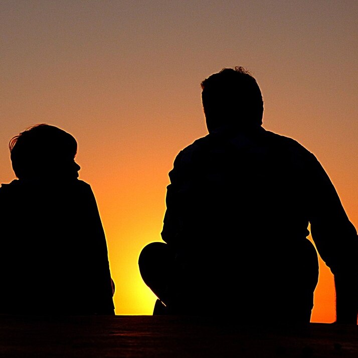 A son and his father are in silhouette facing the sunset.