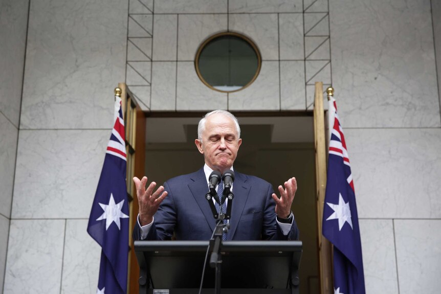 Malcolm Turnbull, flanked by Australian flags, appears to shrug while standing behind a podium at a press conference.