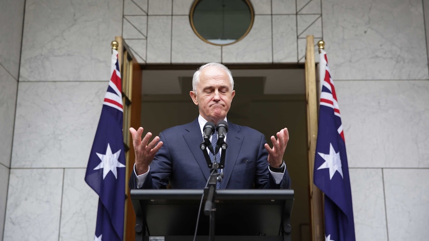 Malcolm Turnbull, flanked by Australian flags, appears to shrug while standing behind a podium at a press conference.