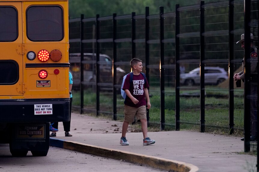 A boy walks from the bus into school gates wearing a shirt that reads 'united we stand'. 