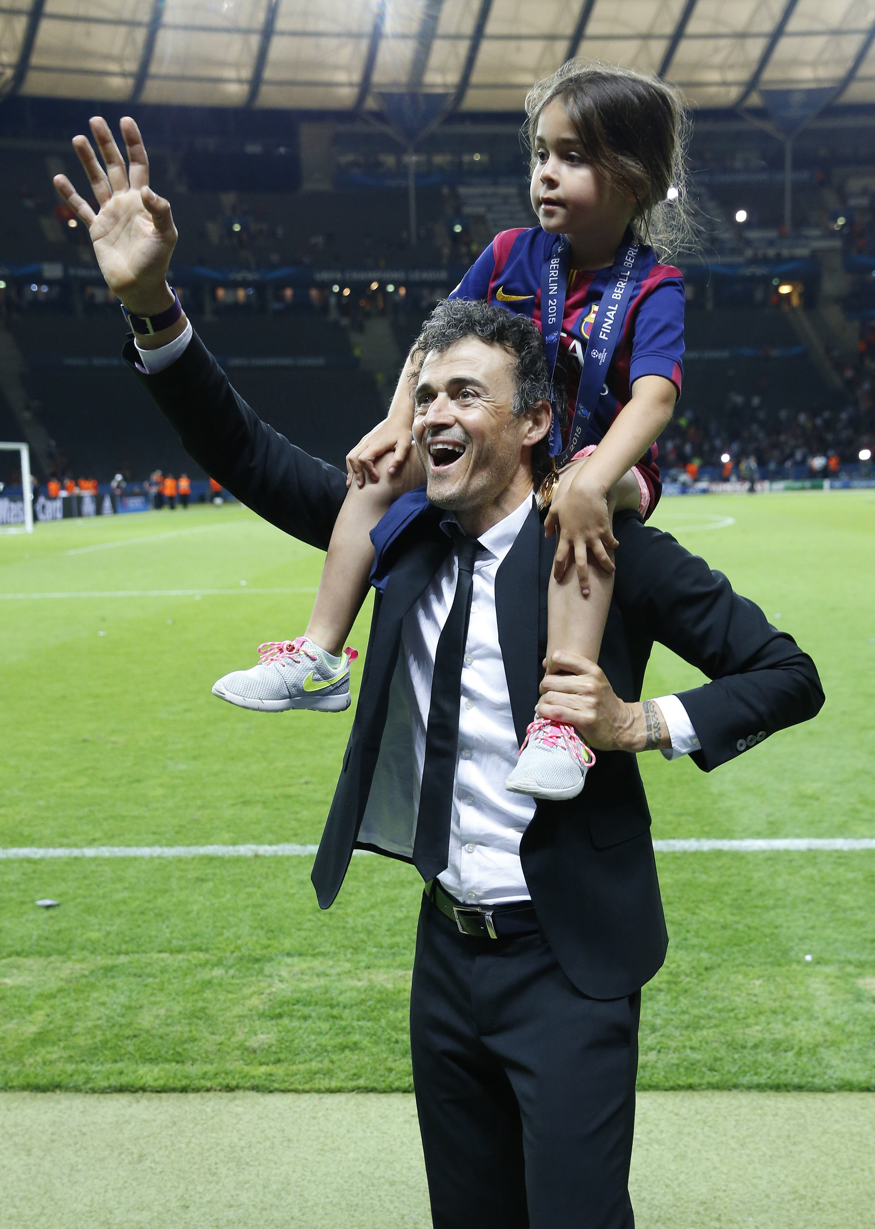 A football coach smiles and waves to the crowd from the pitch after a big match while carrying his daughter on his shoulders.