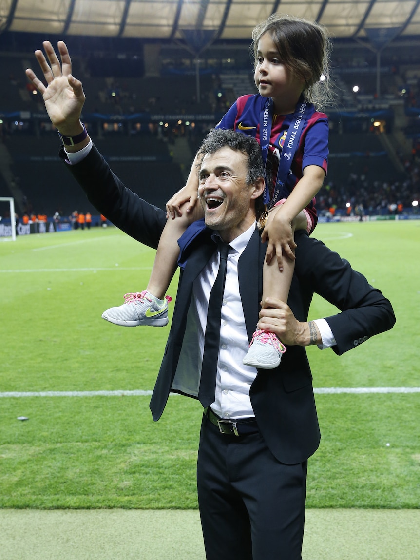 A football coach smiles and waves to the crowd from the pitch after a big match while carrying his daughter on his shoulders.