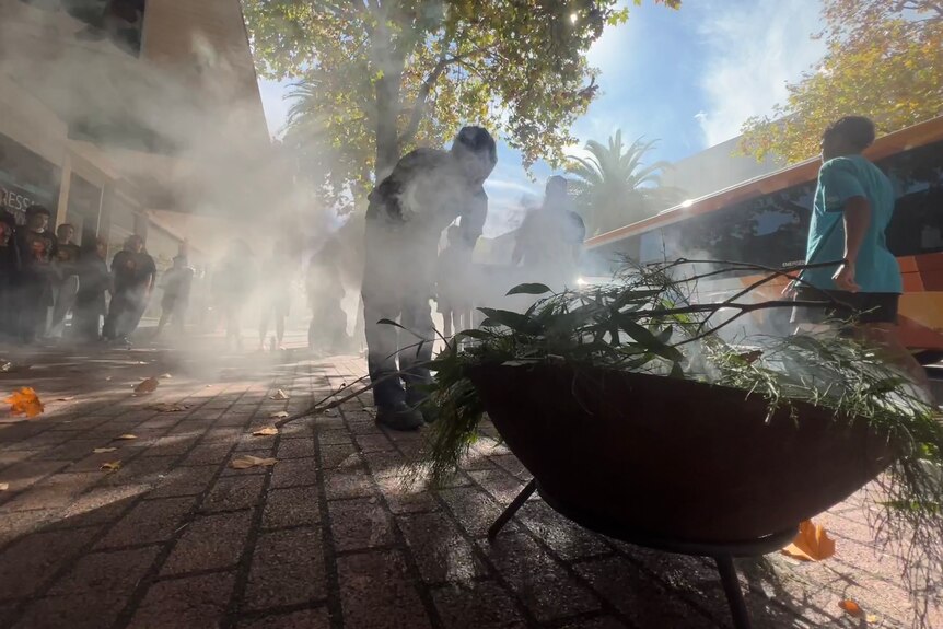 Smoke rises from a large vessel of leaves as part of a ceremony in a suburban setting